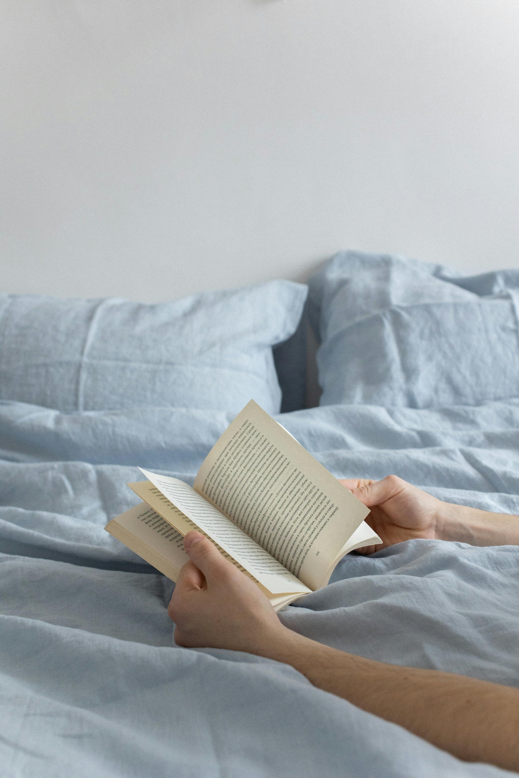 Blue linen bedding set with stacked pillows, cozy blanket, open magazine, and rustic bedside lamp