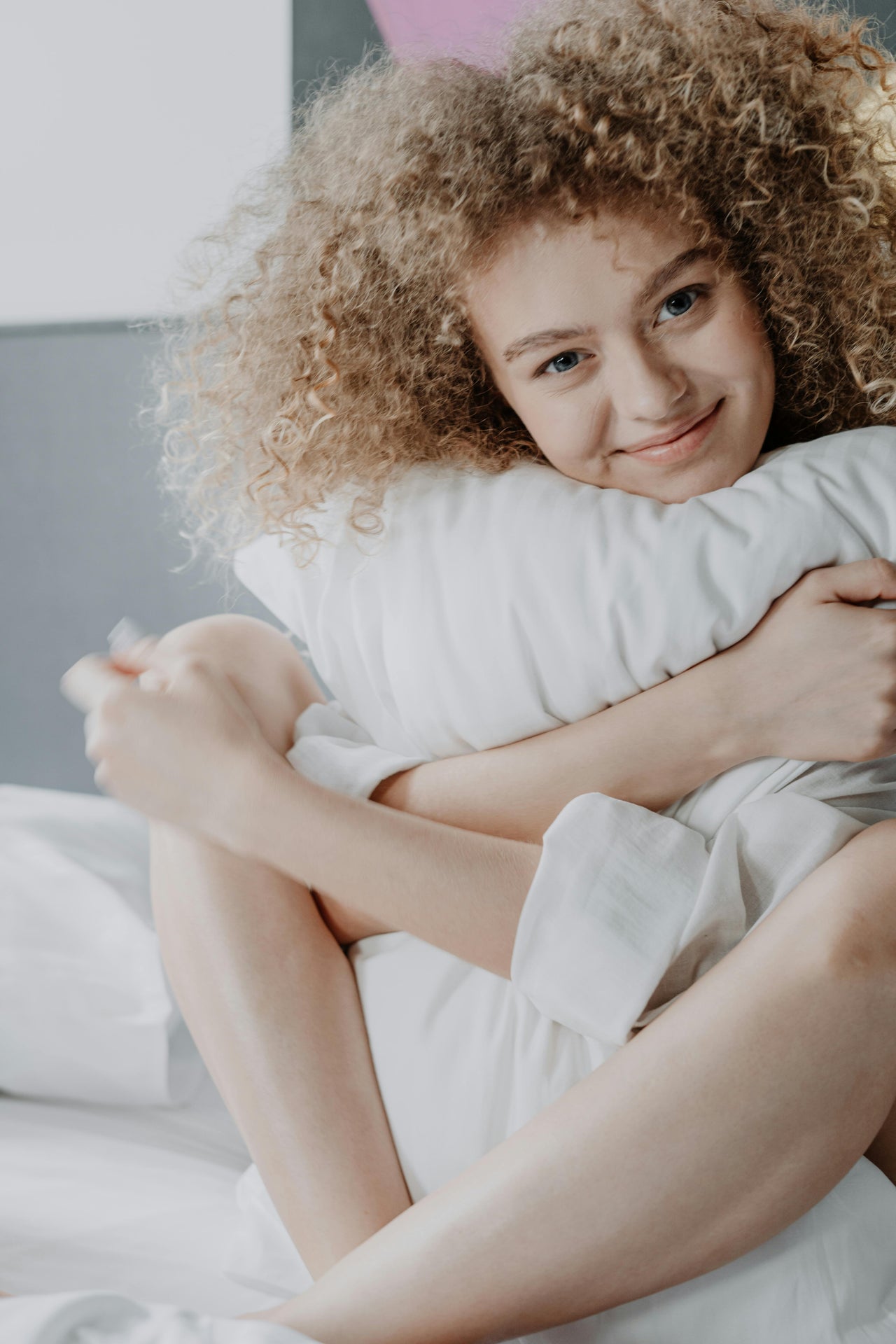 Smiling woman with curly hair hugging a white pillow while sitting on a bed in a cozy bedroom