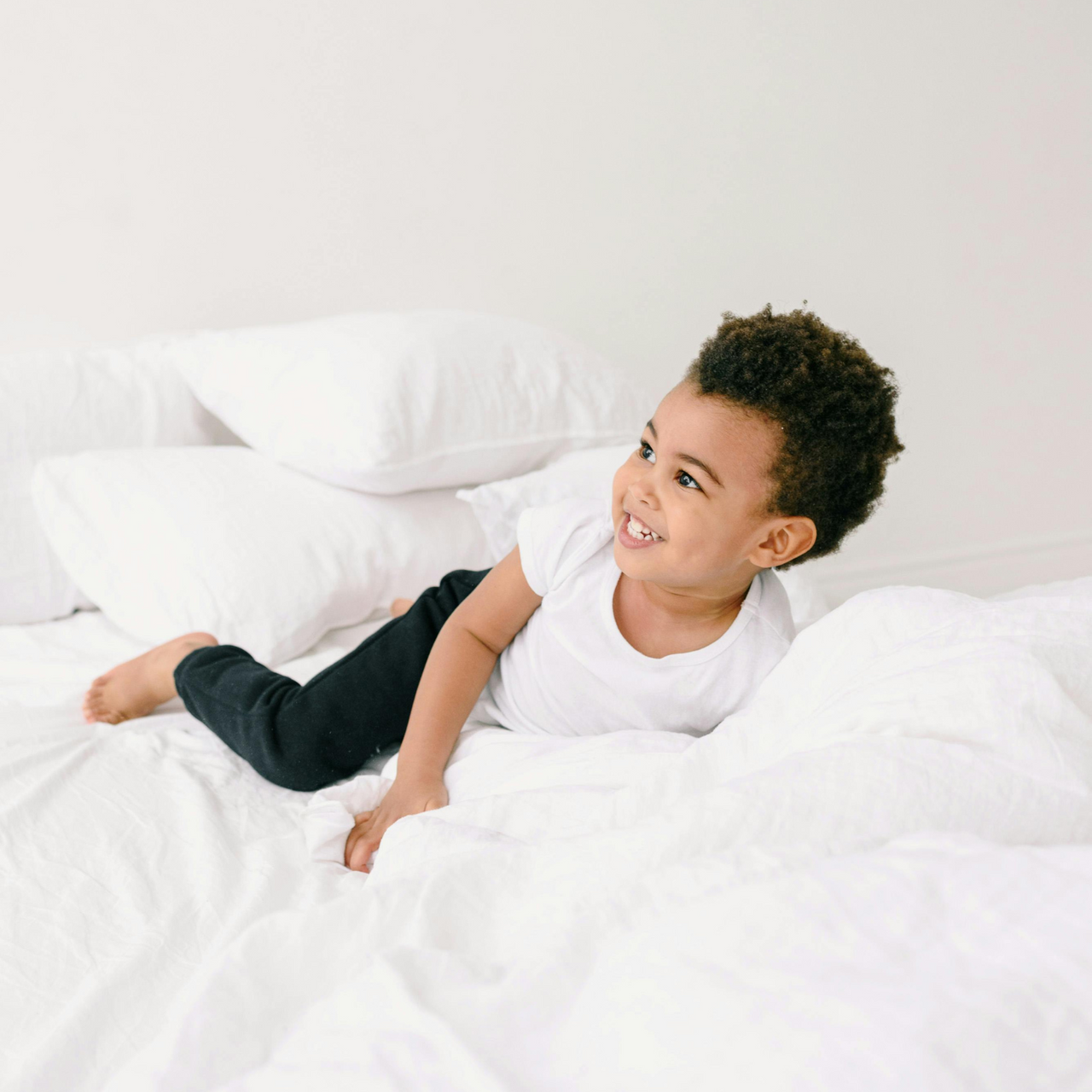 Smiling toddler in white shirt and black pants lying on a white bed with pillows