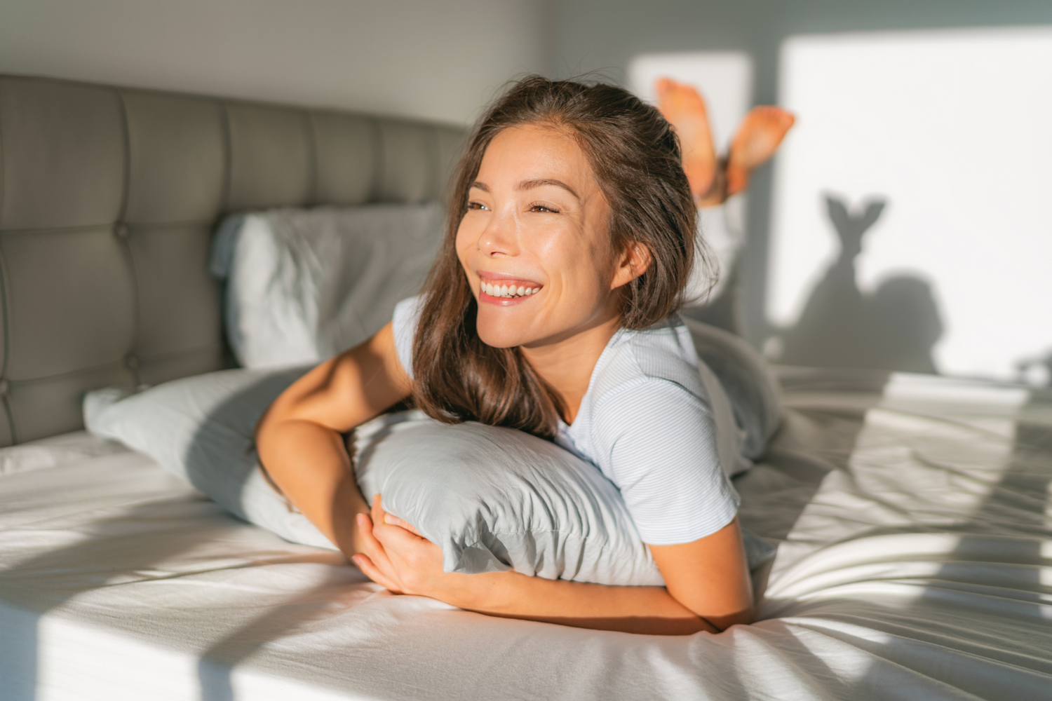 Smiling woman lying on bed hugging pillow in bright sunlight bedroom