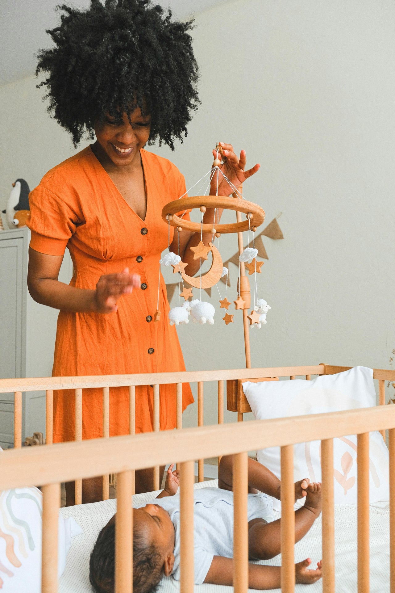Smiling woman in orange dress holding wooden baby mobile above crib with infant in light nursery