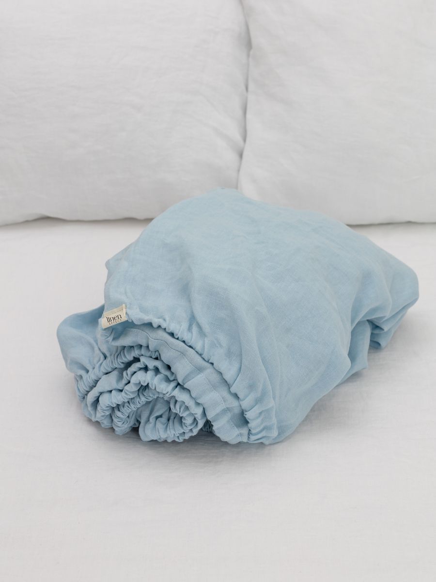 Rolled light blue flax linen fitted sheet on a white bed with white pillows